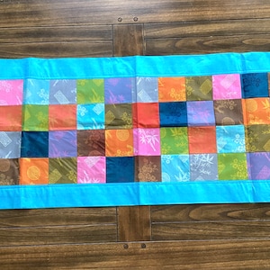 May include: A blue table runner with a patchwork design of colorful squares. The squares feature various patterns, including floral, geometric, and abstract designs.