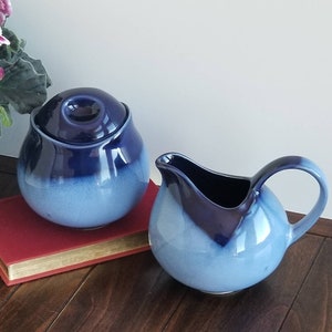May include: A blue and white ceramic creamer and sugar bowl set. The creamer has a handle and the sugar bowl has a lid. Both pieces have a gradient design with a darker blue top and a lighter blue bottom.