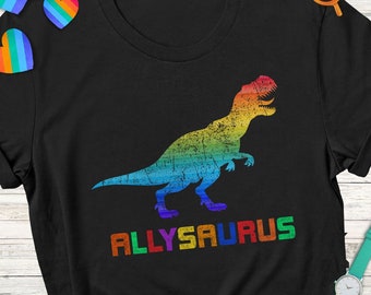 May include: Black t-shirt featuring a rainbow-colored dinosaur graphic with the text "ALLYSAURUS". The design has a distressed, vintage look. The shirt is a crew neck style.