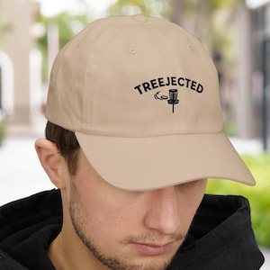 May include: A tan baseball cap with the word "TREEJECTED" in black script above a black disc golf basket graphic. The cap has a curved brim and is worn by a person wearing a black hooded sweatshirt.