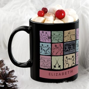 May include: A black ceramic mug with a handle, decorated with colorful fencing illustrations. The mug is filled with marshmallows and red berries. The name "ELIZABETH" is printed on a pink band at the bottom of the mug.