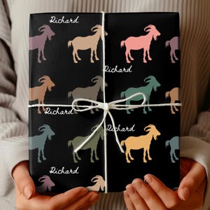 May include: Black gift wrap featuring colorful goat illustrations and the name "Richard" in white script. The gift is tied with white twine. The goats are in shades of pink, tan, green, and blue.