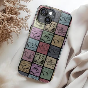 May include: A smartphone case featuring a colorful, geometric design of squares with illustrations of a cornhole game. The case has a clear border and a multi-camera lens cutout. The background is a white, silky fabric.