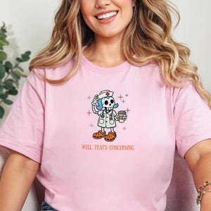 May include: A light pink t-shirt featuring a graphic of a skeleton dressed as a nurse with blue hair, wearing a stethoscope and holding a coffee cup. The text "WELL THAT'S CONCERNING" is printed in orange below the graphic. The shirt has short sleeves.