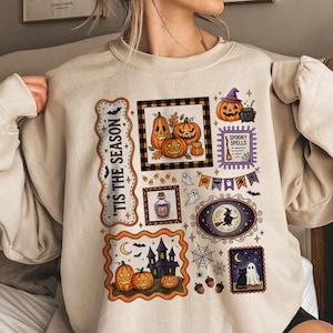 Könnte beinhalten: Ein sandfarbenes Sweatshirt mit Rundhalsausschnitt und einem mehrteiligen Grafikdruck mit Halloween-Motiven wie Kürbissen, einem Spukhaus, einer schwarzen Katze, einem Geist, einer Hexe auf einem Besen sowie dem Text 'TIS THE SEASON' und 'SPOOKY SPELLS'.