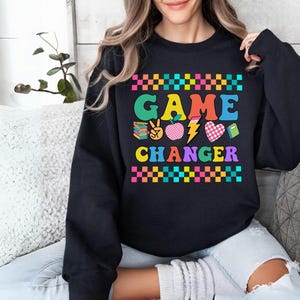 May include: Black sweatshirt with the words "GAME CHANGER" in colorful block letters. The design includes a peace sign, apple, lightning bolt, and heart. The top and bottom of the text are bordered with a checkered pattern.