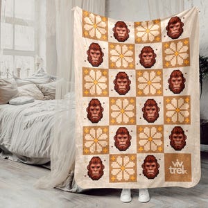 May include: A beige blanket with a repeating pattern of pixelated ape faces and floral designs in brown and yellow. The blanket is held up by a person wearing white shoes. The blanket has the text "viv trek" in the bottom right corner.