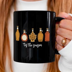 May include: Black ceramic mug featuring a series of illustrated bottles in shades of brown and tan. The phrase "tis the season" is written in white script below the bottles. The mug has a black handle.