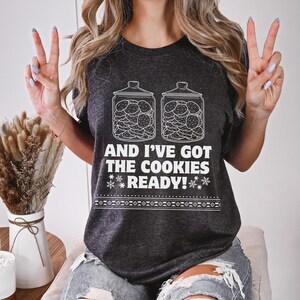 K&ouml;nnte beinhalten: Ein dunkelgraues T-Shirt mit einer wei&szlig;en Grafik von zwei mit Keksen gef&uuml;llten Keksdosen. Der Text auf dem Shirt lautet "And I've got the cookies ready!"