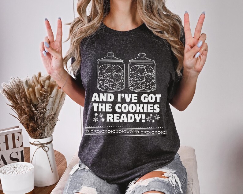 K&ouml;nnte beinhalten: Ein dunkelgraues T-Shirt mit einer wei&szlig;en Grafik von zwei mit Keksen gef&uuml;llten Keksdosen. Der Text auf dem Shirt lautet "And I've got the cookies ready!"