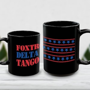 Könnte beinhalten: Zwei schwarze Keramikbecher. Der linke Becher zeigt den roten und blauen Schriftzug "FOXTROT DELTA TANGO" in gestapelter Anordnung. Der rechte Becher zeigt ein wiederkehrendes Muster aus horizontalen roten Linien und Reihen blauer Sterne.