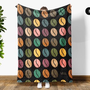 May include: A black throw blanket featuring a repeating pattern of coffee beans in various muted colors including yellow, green, blue, orange, and pink. The blanket has a dark background with a subtle star-like texture and a small logo in the corner.