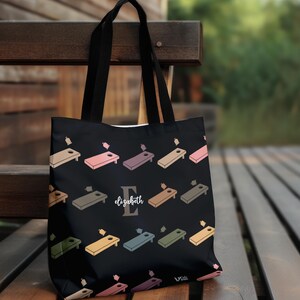 May include: A black tote bag featuring a repeating pattern of colorful cornhole boards and bean bags. The center of the bag displays a large grey letter E with the name elizabeth written in white script font across the letter.