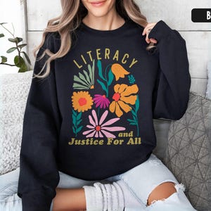 May include: Black sweatshirt with a floral design and the words "Literacy and Justice For All" in gold lettering. The floral design includes orange, pink, and yellow flowers with green leaves. The sweatshirt is a crew neck style.