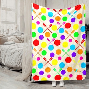 May include: A colorful blanket with a pattern of large, multi-colored polka dots and crossed oars. The dots are red, orange, yellow, green, blue, and purple. The oars are red, green, and brown. The blanket is held up by a person wearing white shoes.