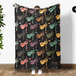 May include: Black blanket featuring a repeating pattern of colorful violins and bows. The violins are in shades of orange, green, purple, and yellow. The blanket is held up by a person, and the background is a wooden floor and a green plant.
