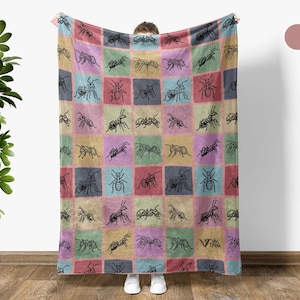 May include: A colorful throw blanket featuring a patchwork design with black ant illustrations on squares of pink, blue, green, and yellow. The blanket is held up, showcasing its design and texture.