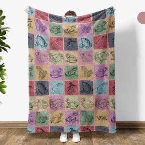 Puede incluir: Una manta de forro polar suave con un patr&oacute;n colorido de ranas en un dise&ntilde;o de cuadr&iacute;cula. La manta es de color rosa apagado con el texto "Old Rose" en un cuadro blanco en la esquina superior derecha.