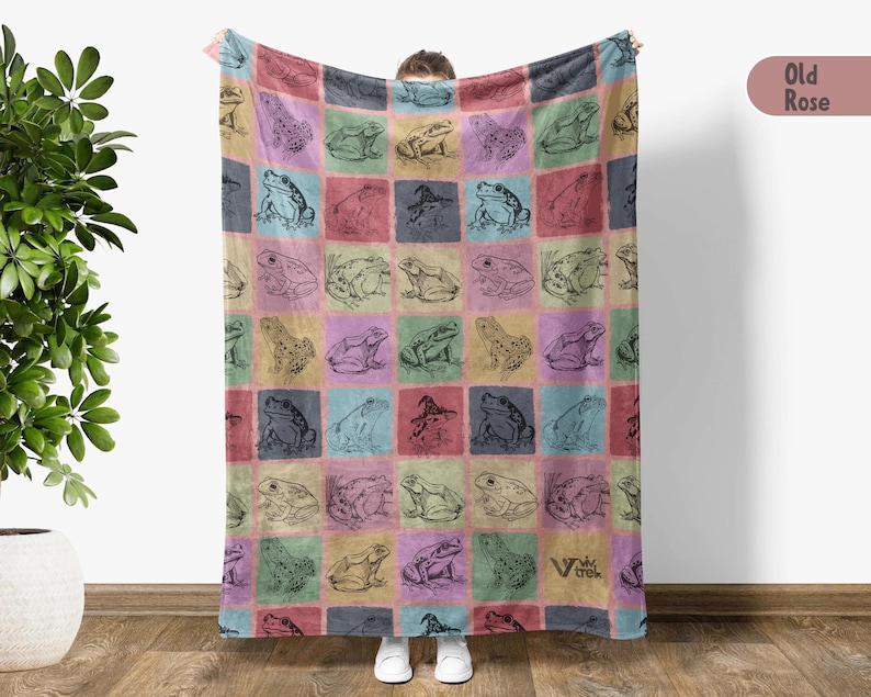 Puede incluir: Una manta de forro polar suave con un patr&oacute;n colorido de ranas en un dise&ntilde;o de cuadr&iacute;cula. La manta es de color rosa apagado con el texto "Old Rose" en un cuadro blanco en la esquina superior derecha.