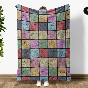 May include: A colorful fleece blanket with a repeating pattern of monkeys in different poses. Each monkey is outlined in black and set against a different colored square. The blanket is held up by a person, but the person's hands are not visible.