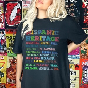 May include: Black t-shirt with the text "Hispanic Heritage" in rainbow colors. The text is followed by a list of countries in South and Central America, including Argentina, Brazil, Chile, Dominican Republic, Ecuador, El Salvador, Guatemala, Puerto Rico, Honduras, Mexico, Cuba, Costa Rica, Nicaragua, Peru, Panama, Uruguay, Bolivia, Paraguay, Spain, Colombia, Venezuela, and USA.
