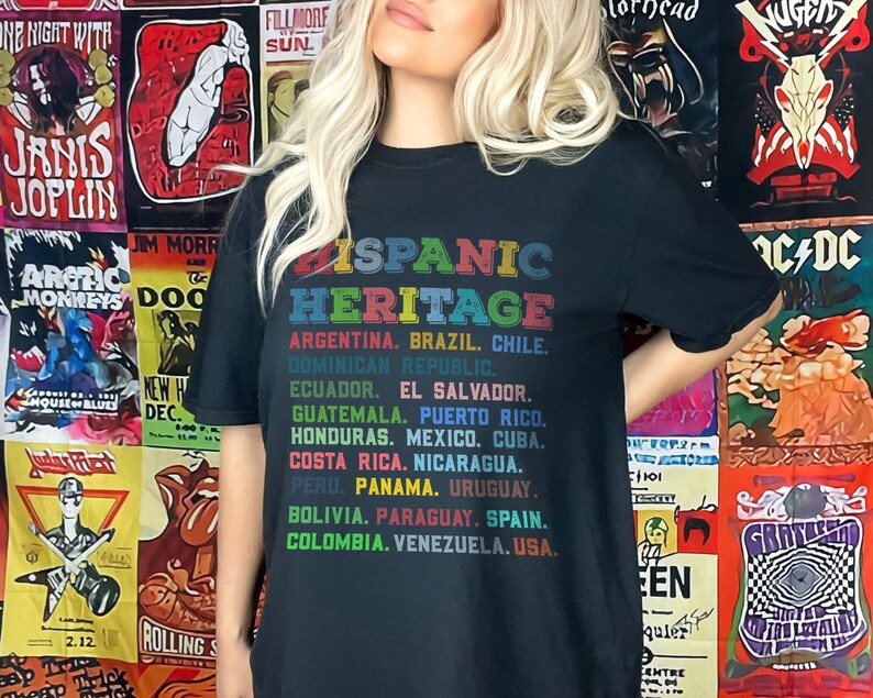 May include: Black t-shirt with the text "Hispanic Heritage" in rainbow colors. The text is followed by a list of countries in South and Central America, including Argentina, Brazil, Chile, Dominican Republic, Ecuador, El Salvador, Guatemala, Puerto Rico, Honduras, Mexico, Cuba, Costa Rica, Nicaragua, Peru, Panama, Uruguay, Bolivia, Paraguay, Spain, Colombia, Venezuela, and USA.