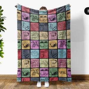 May include: A black throw blanket featuring a repeating grid pattern of colorful squares. Each square contains black line art illustrations of coffee-related items including espresso machines, moka pots, coffee cups, beans, and croissants.