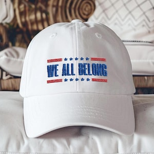 May include: White baseball cap with blue embroidery that reads "We All Belong" with red and white stars above and below the text.