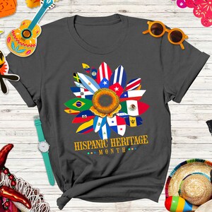 May include: A dark gray t-shirt with a sunflower design made up of flags from different Hispanic countries. The text "HISPANIC HERITAGE MONTH" is printed below the sunflower.