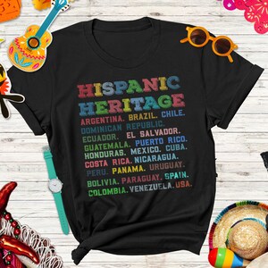 May include: Black t-shirt with a colorful graphic print listing countries in the Hispanic Heritage. The text reads "HISPANIC HERITAGE" and lists countries including Argentina, Brazil, Chile, Dominican Republic, Ecuador, El Salvador, Guatemala, Puerto Rico, Honduras, Mexico, Cuba, Costa Rica, Nicaragua, Peru, Panama, Uruguay, Bolivia, Paraguay, Spain, Colombia, Venezuela, and USA.