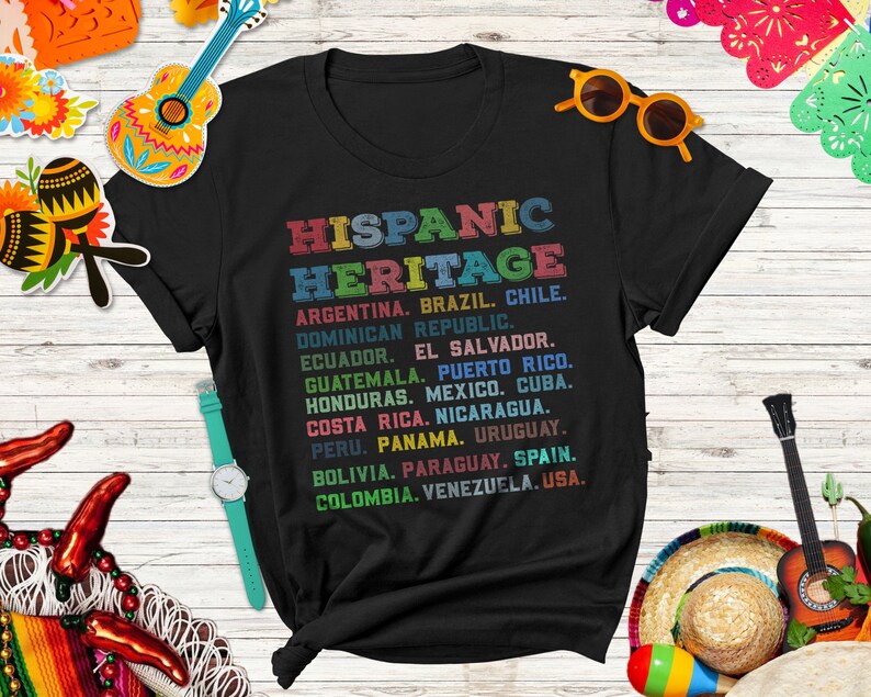May include: Black t-shirt with a colorful graphic print listing countries in the Hispanic Heritage. The text reads "HISPANIC HERITAGE" and lists countries including Argentina, Brazil, Chile, Dominican Republic, Ecuador, El Salvador, Guatemala, Puerto Rico, Honduras, Mexico, Cuba, Costa Rica, Nicaragua, Peru, Panama, Uruguay, Bolivia, Paraguay, Spain, Colombia, Venezuela, and USA.