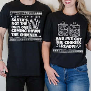 K&ouml;nnte beinhalten: Zwei schwarze T-Shirts mit wei&szlig;em Text und Grafiken. Das erste Shirt sagt "Santa's not the only one coming down the chimney..." Das zweite Shirt sagt "And I've got the cookies ready!" mit einer Grafik von zwei Keksdosen.