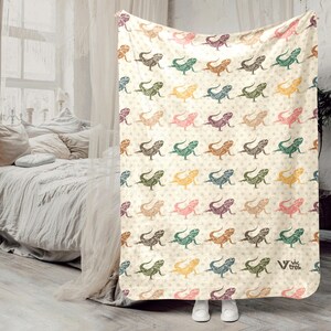 May include: A white throw blanket with a repeating pattern of colorful lizards. The blanket has a cream background with a subtle floral pattern. The lizards are in various shades of green, blue, orange, and pink. The blanket has the text "V-Trek" in the bottom right corner.