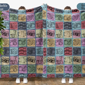 May include: A large, colorful blanket featuring a repeating pattern of vintage cameras in various colors, including pink, blue, yellow, and green. The design also includes film strips. The blanket is held up by two people.