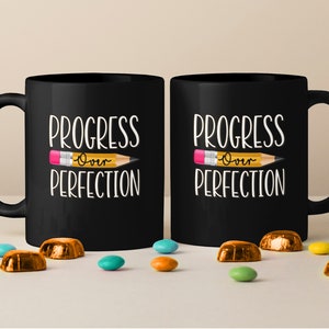 Könnte beinhalten: Zwei schwarze Keramikbecher mit dem weißen Text "PROGRESS" und "PERFECTION" sowie einer gelben Bleistiftgrafik in der Mitte, die das Wort "Over" in Schreibschrift enthält. Die Becher haben einen Standardhenkel und eine glatte, matte Oberfläche.