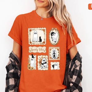 Könnte beinhalten: Ein orangefarbenes T-Shirt mit einem Grafikdruck aus mehreren rechteckigen und ovalen Rahmen, die Illustrationen von weißen Geistern und schwarzen Katzen enthalten, mit herbstlichen Blumenakzenten und dem Text Spooky Vibes in der Mitte.
