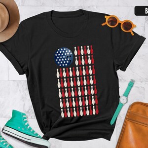 Może przedstawiać: Czarny t-shirt z grafiką przedstawiającą amerykańską flagę złożoną z kręgli i kuli do kręgli. Kula jest niebieska w białe gwiazdy, a pasy tworzą naprzemienne rzędy czerwonych i białych kręgli ułożonych w kształt flagi Stanów Zjednoczonych.