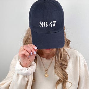 Może przedstawiać: Granatowa czapka z daszkiem z białymi cyframi "8647" z przodu. Czapka ma zakrzywiony daszek i klasyczny sześciopanelowy design. Swobodny dodatek do noszenia na co dzień.