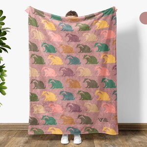 May include: A rectangular throw blanket in an old rose color featuring a repeating pattern of stylized badgers in various colors including yellow, teal, green, and purple. The blanket includes a small logo in the bottom right corner.