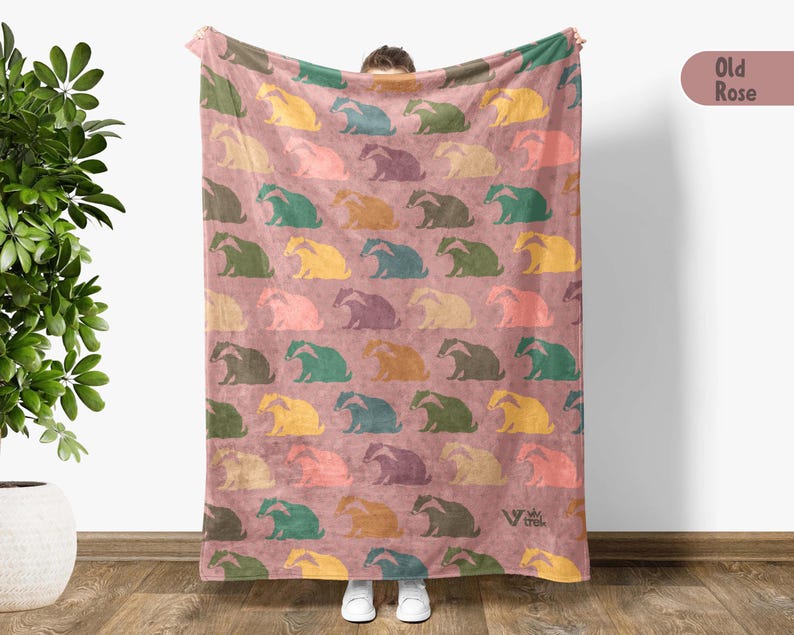 May include: A rectangular throw blanket in an old rose color featuring a repeating pattern of stylized badgers in various colors including yellow, teal, green, and purple. The blanket includes a small logo in the bottom right corner.