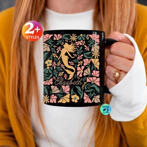 Könnte beinhalten: Ein schwarzer Keramikbecher mit einer goldenen Meerjungfrauen-Silhouette, umgeben von einem Muster aus rosa, gelben und grünen Blumen und Blättern. Der Name Elizabeth ist in weißer Schreibschrift mittig aufgedruckt, darunter steht vivtek.