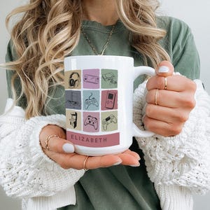 May include: White ceramic mug featuring a colorful grid of gaming-related icons, including headphones, game consoles, and controllers. The name "ELIZABETH" is printed in a burgundy band at the bottom. The mug has a large handle and is held by a person.