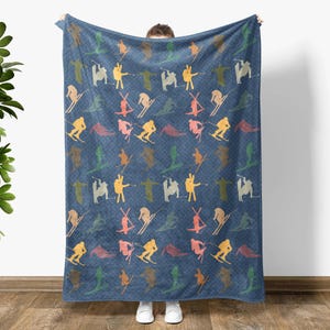 May include: A blue fleece blanket with a repeating pattern of colorful skiers on a white background. The blanket is being held up by a person.