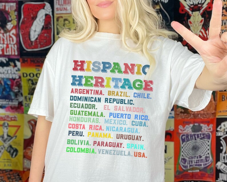 May include: White t-shirt with the text "HISPANIC HERITAGE" in rainbow colors. The text is followed by a list of countries in Spanish-speaking regions, including Argentina, Brazil, Chile, Dominican Republic, Ecuador, El Salvador, Guatemala, Puerto Rico, Honduras, Mexico, Cuba, Costa Rica, Nicaragua, Peru, Panama, Uruguay, Bolivia, Paraguay, Spain, Colombia, Venezuela, and USA.