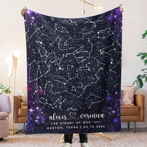 May include: A dark blue blanket featuring a white constellation design with a purple starry border. The blanket has text that reads "alexis veronica THE STRART OF OUR US HUSTON, TEXAS 05 10 2024". The blanket is held up in front of a living room.