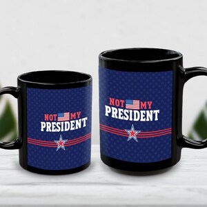 Könnte beinhalten: Zwei schwarze Keramiktassen mit blauem Hintergrund und dezentem Sternenmuster. Der Text NOT MY PRESIDENT ist in Weiß mit einer kleinen amerikanischen Flagge aufgedruckt. Unter dem Text befinden sich drei rote Linien und ein roter Stern.