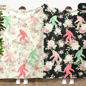 May include: Cream white and black blankets with a floral and Bigfoot pattern. The cream white blanket has a pattern of pink, green, and cream-colored Bigfoot figures and flowers. The black blanket has a pattern of pink and green Bigfoot figures and flowers.