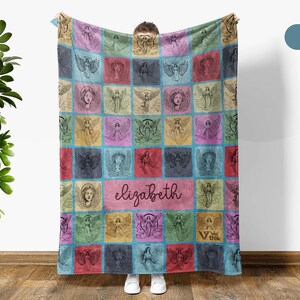 May include: A rectangular throw blanket featuring a grid of colorful squares, each displaying a unique black line-art illustration of an angel. The center square contains the name elizabeth in cursive script, and the logo viv trek is visible.