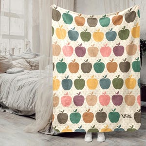 May include: A cream-colored blanket with a repeating pattern of colorful apples in shades of green, yellow, orange, pink, and purple. The blanket is draped over a bed, with the feet of a person wearing white sneakers visible at the bottom.
