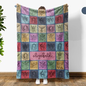 May include: A rectangular throw blanket featuring a grid of colorful squares, each containing a black line-art illustration of an angel. A central horizontal rectangle displays the name elizabeth in cursive script. The blanket has a marble blue color.